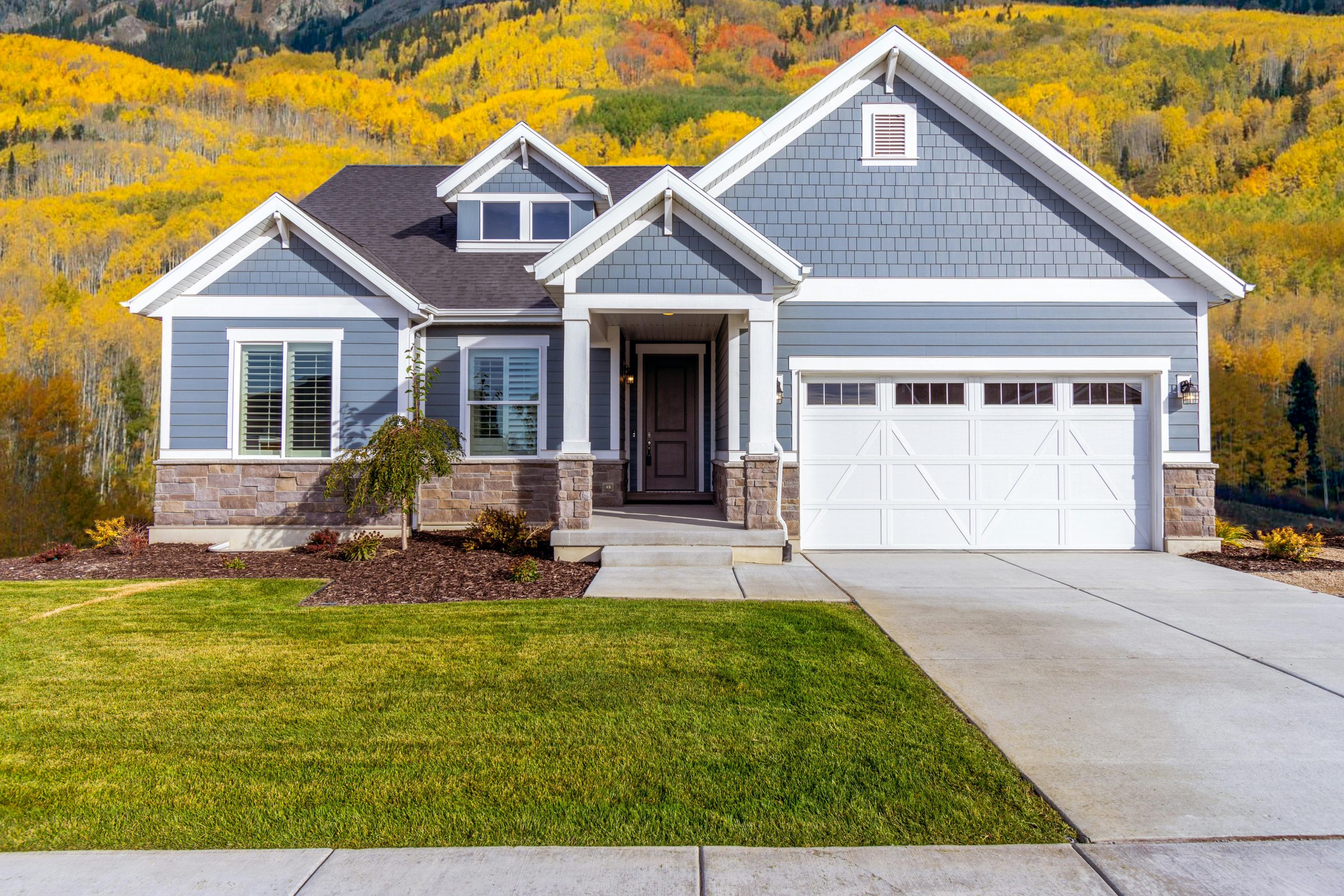 Front view of a blue house in a mountain neighborhood, surrounded by trees with autumn-colored leaves, used to indicate now is a good time to sell your home.